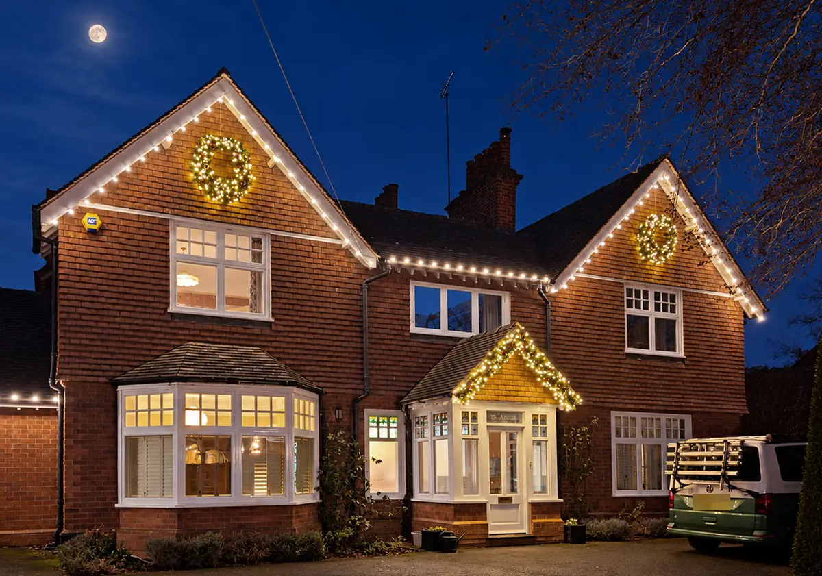 Brick house at dusk with warm white roofline lights, lit wreaths on both gables and a lit porch