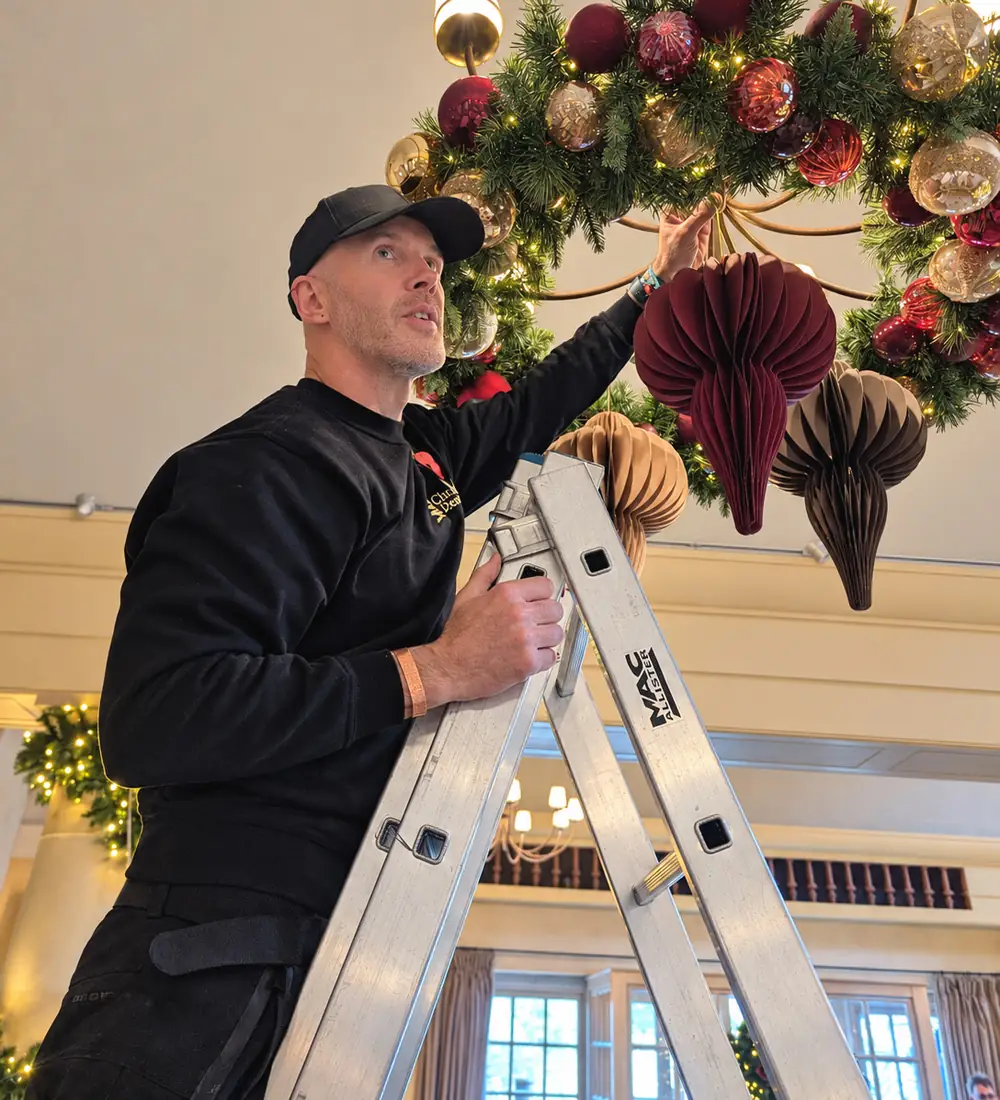 Gareth up a ladder dressing a chandelier with a garland and baubles
